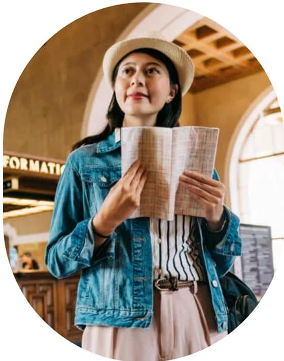 A woman in a straw hat and denim jacket reading a folded map in a station concourse, beneath an information sign.