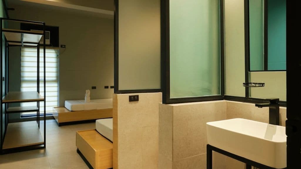 A room with an open en-suite: a white rectangular basin on a black steel frame, a green glass partition, and two low timber-plinth beds beyond.