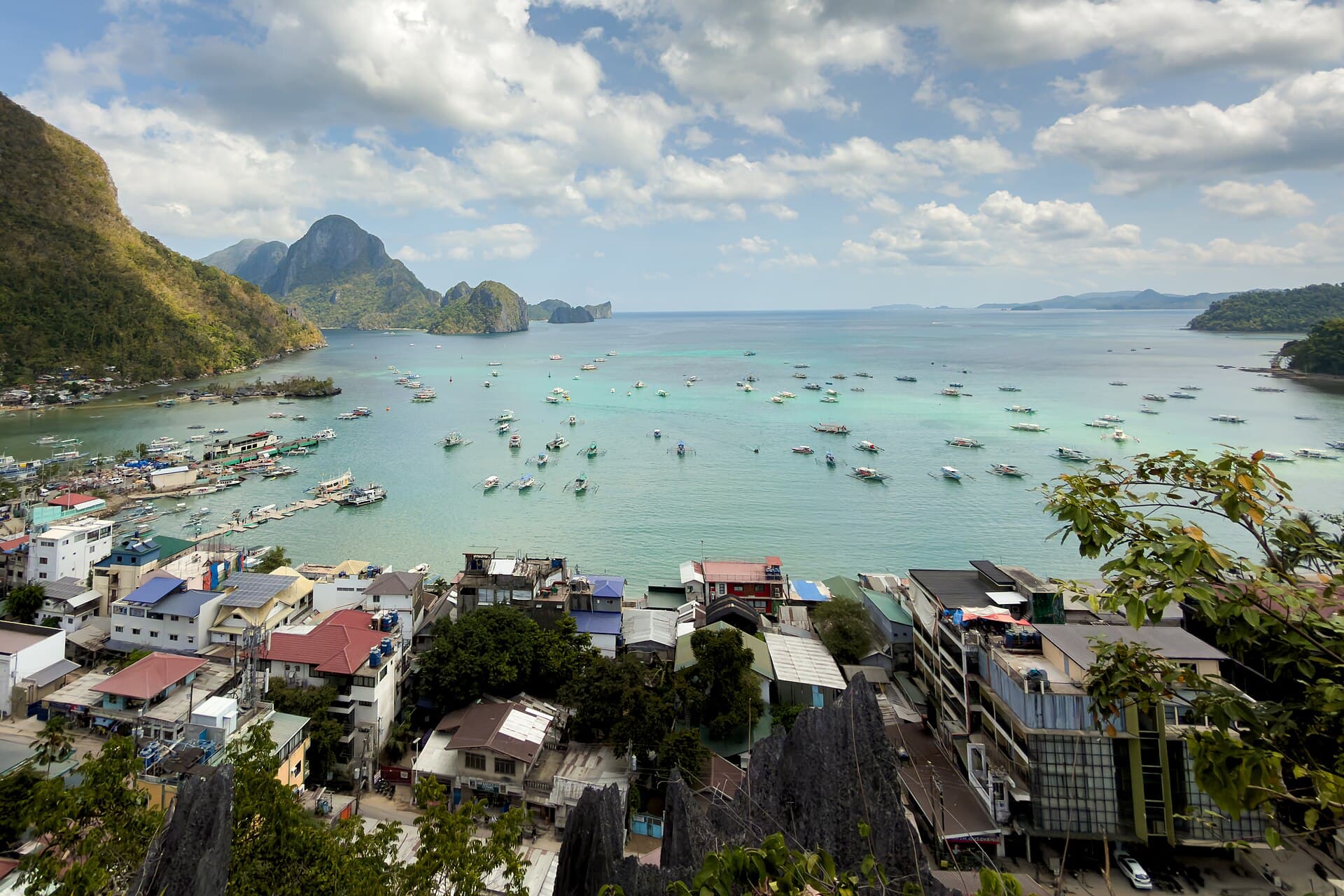 El Nido town and Bacuit Bay seen from above, dozens of white outrigger boats moored across turquoise shallows with karst islands on the horizon.