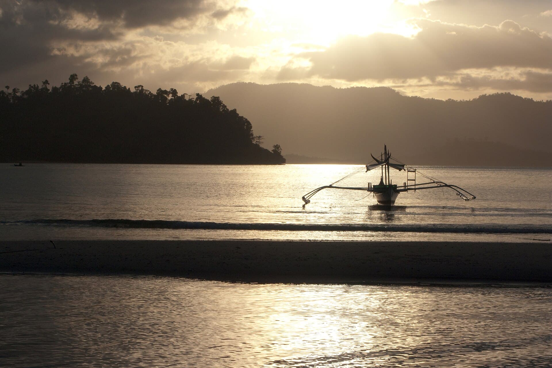 An outrigger bangka silhouetted on calm water at Port Barton, a low sun breaking through cloud behind a forested headland.