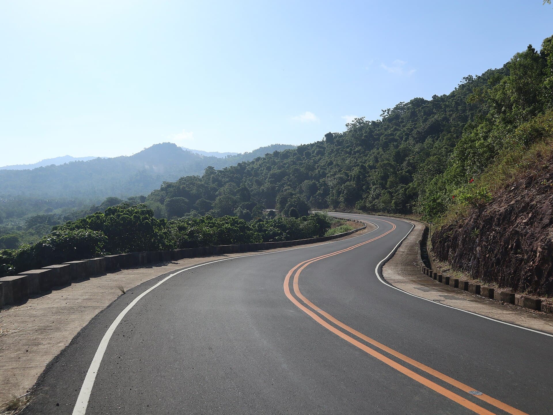 The Taytay to El Nido highway curving away through forested hills, double orange centre lines on fresh asphalt and layered ridges fading into haze.
