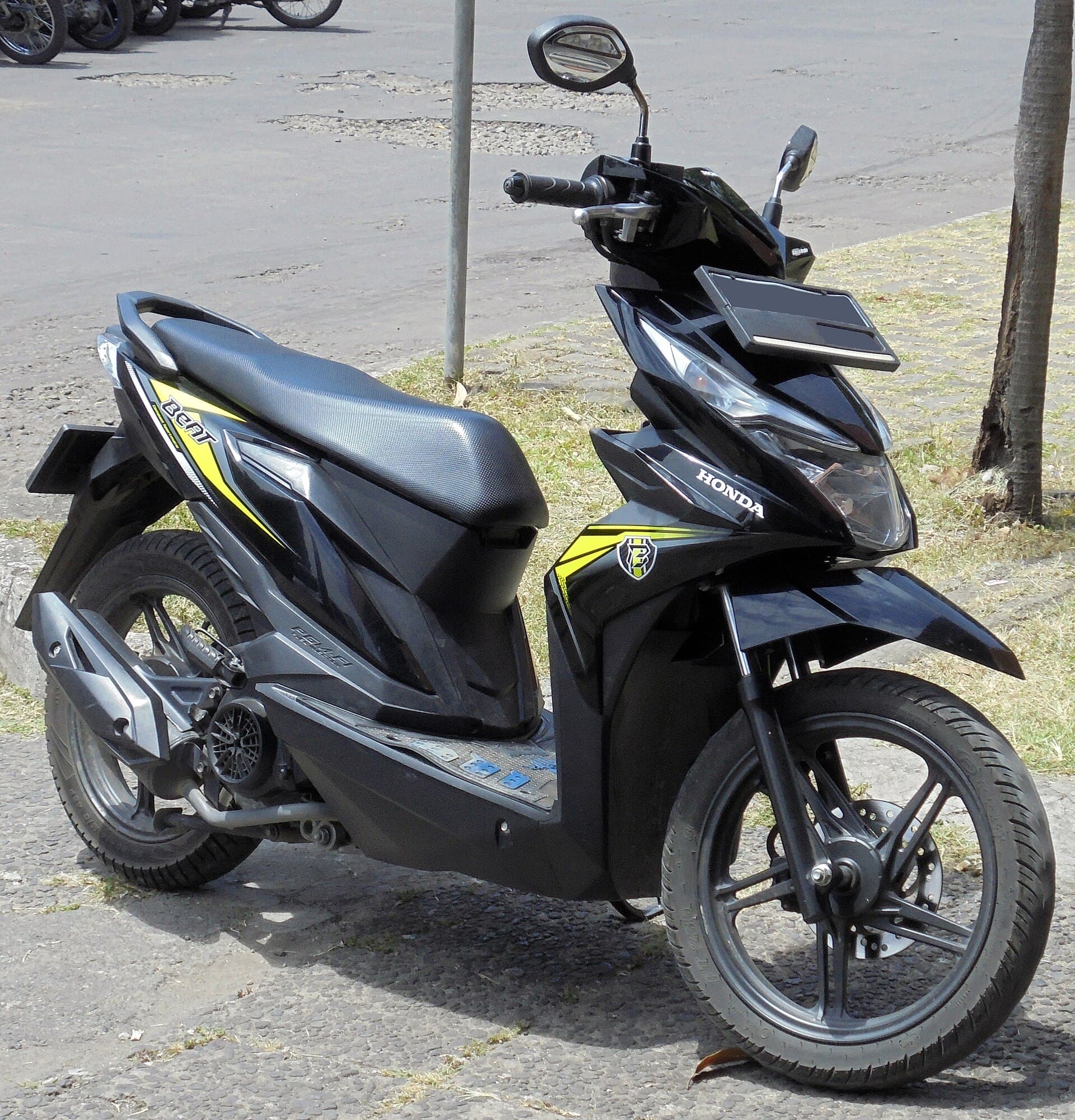 A black Honda BeAT scooter with lime-yellow graphics parked on a kerbside grass strip, front three-quarter view, a palm trunk behind.