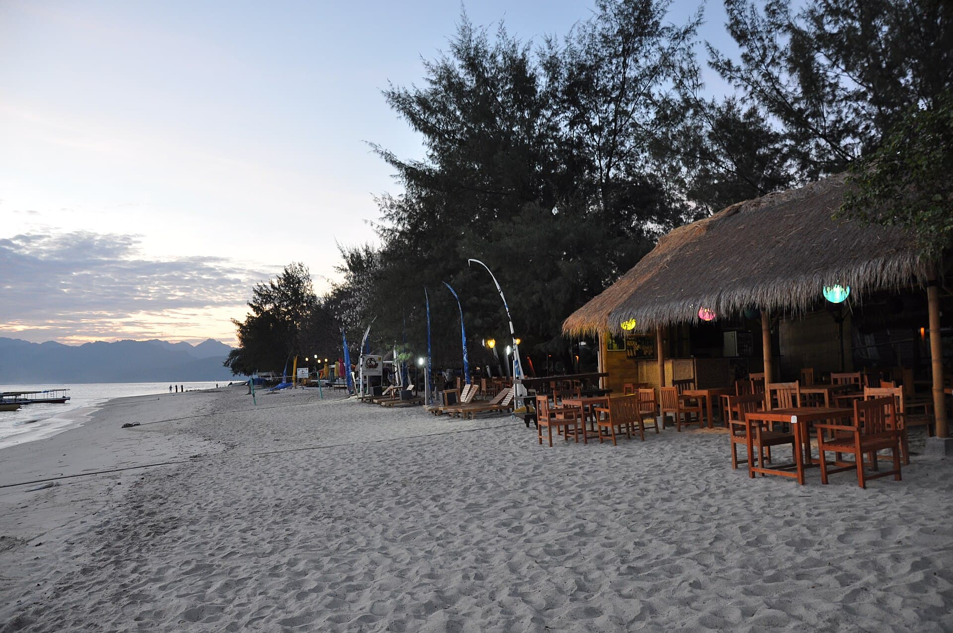 A thatched beachfront bar at dusk with timber tables set out on pale sand, casuarina trees behind and coloured lanterns under the eaves.
