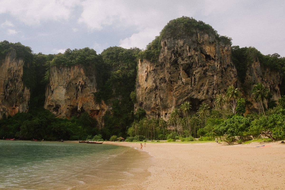 A wide sand beach below towering limestone cliffs with jungle on their ledges, a thatched roof among the palms and boats moored along the shore.