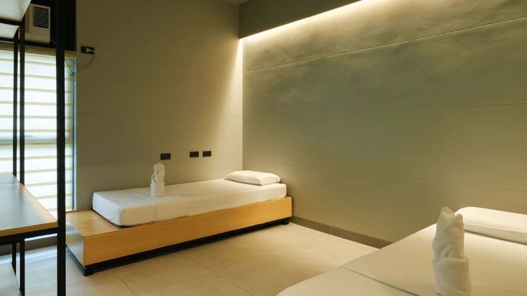 A twin room with two low beds on pale timber plinths, white linen and folded towels, warm cove lighting along a sage rendered wall.
