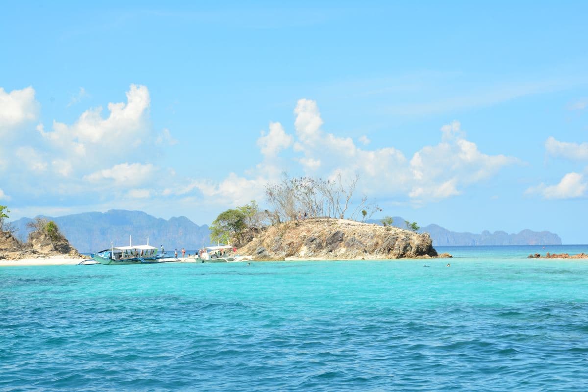 A small rocky islet across banded turquoise water, outrigger bangka boats moored at its sand spit and a range of blue karst islands on the horizon.