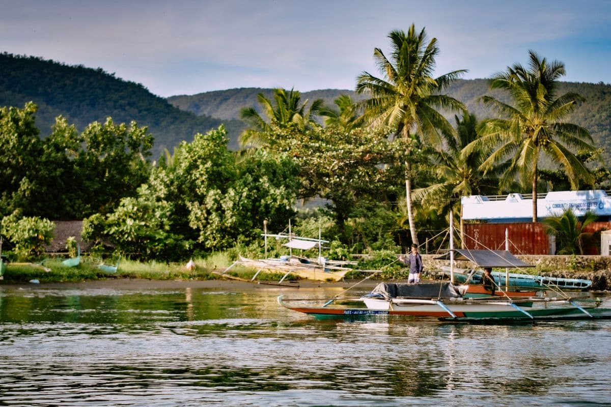 An outrigger bangka moored in calm green water at a village landing, a man standing on the bow, coconut palms and a corrugated shed behind.