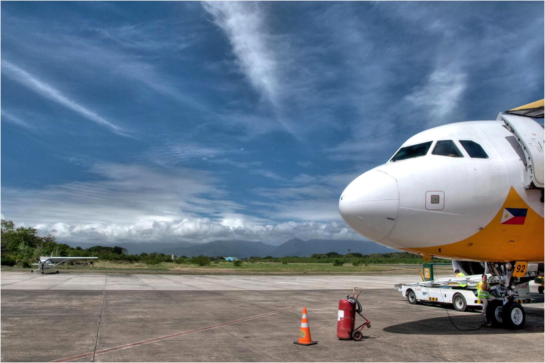 The nose of a white and yellow airliner on the apron at Puerto Princesa, ground crew in hi-vis vests beside the nose gear, blue mountains and towering cloud behind.