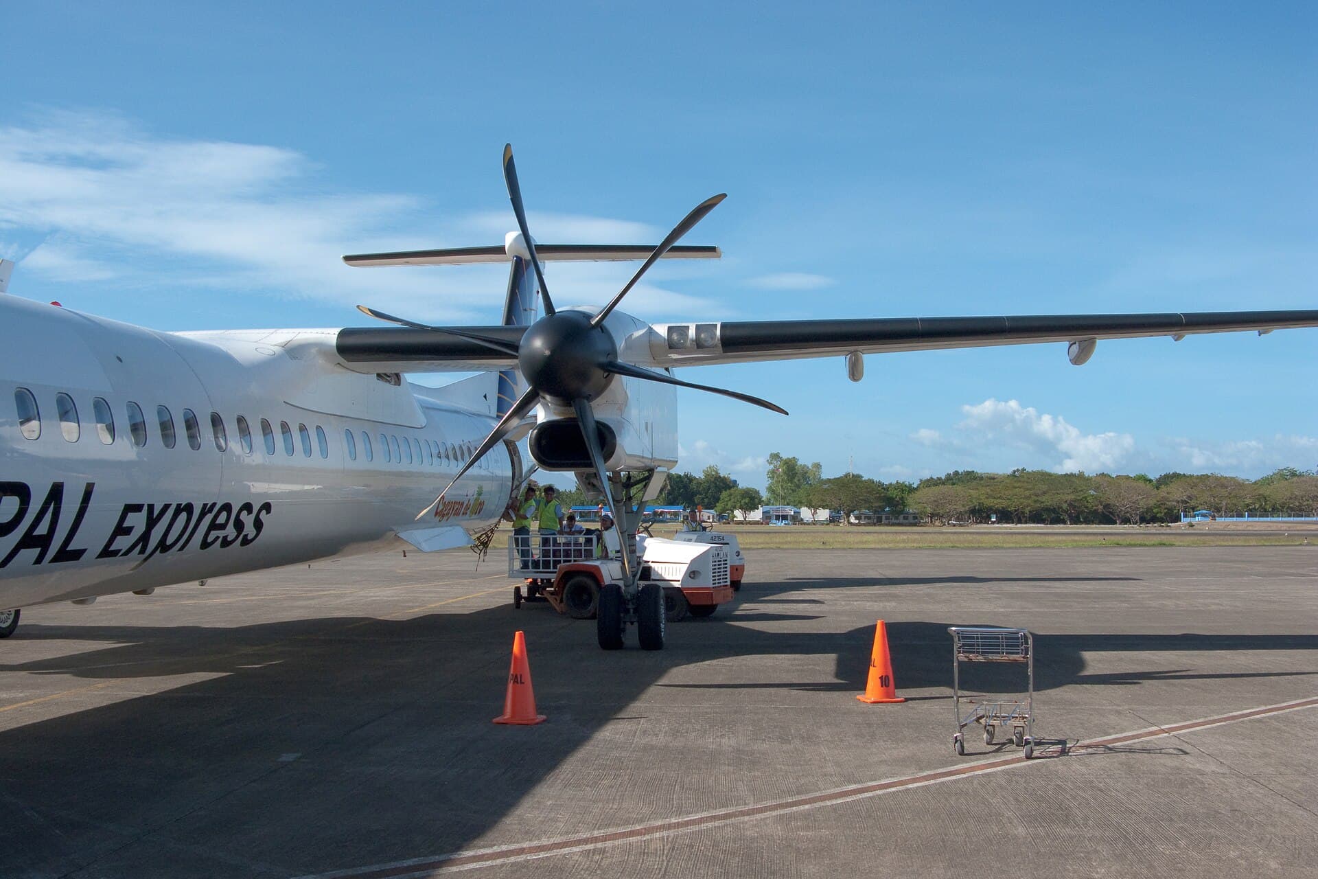 A white turboprop airliner on the apron with its six-bladed propeller in the foreground, passengers at the airstairs and baggage carts alongside.