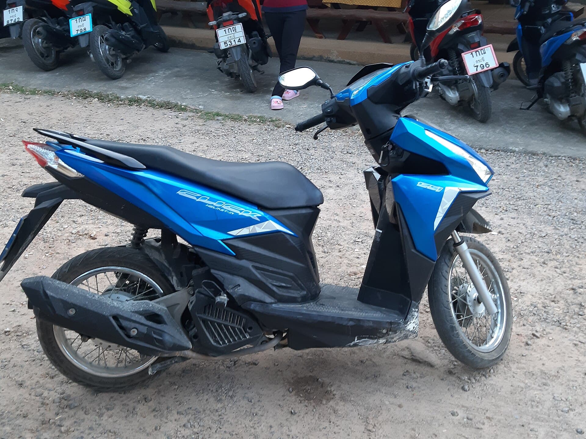 A blue and black Honda Click 125 automatic scooter parked on gravel, seen from its left side, with a rank of other scooters behind.