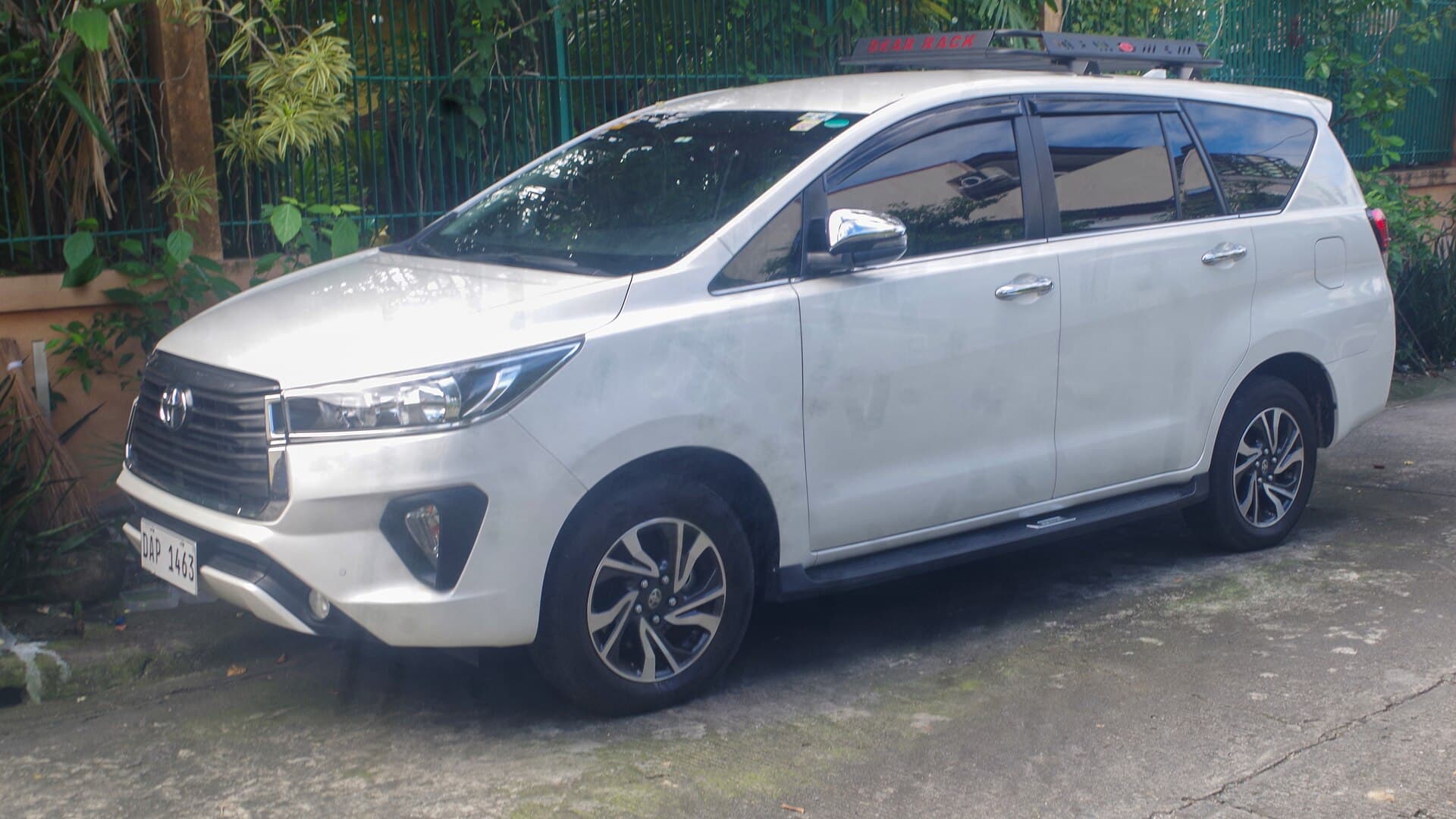 A white Toyota Innova seven-seat van with a roof rack, parked on a concrete driveway against a green fence and tropical foliage.