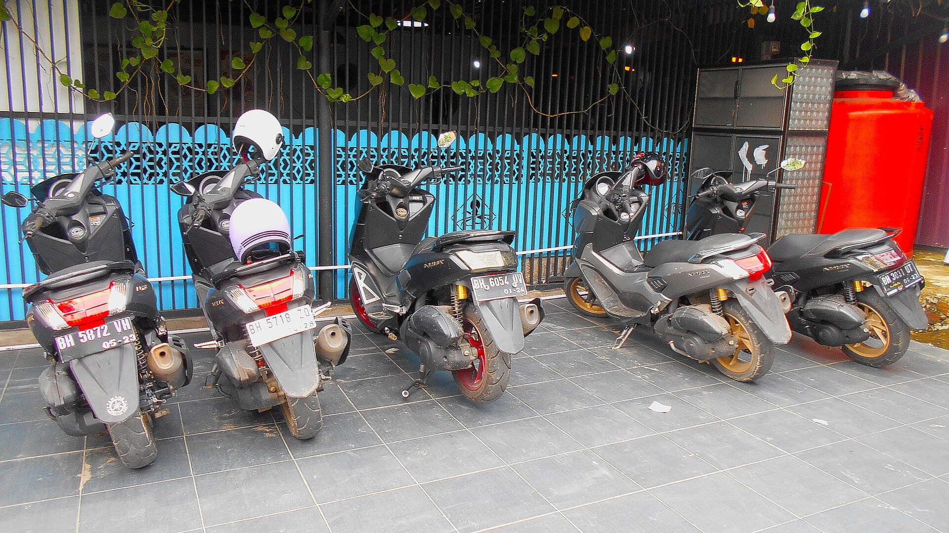 Six black Yamaha NMAX scooters parked in a row against a bright blue fence, two open-face helmets hooked on the mirrors.