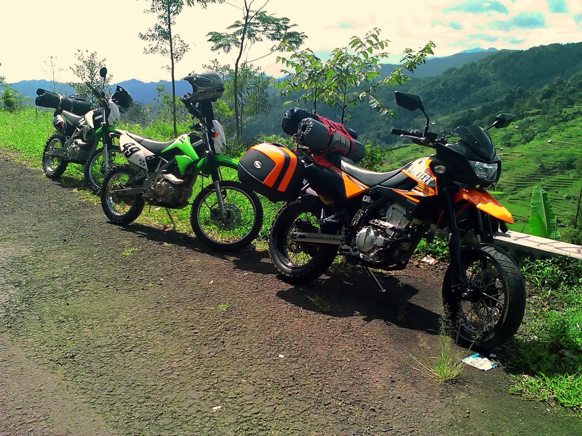 Three 150cc trail bikes parked on a gravel track above green terraced hillsides, bags strapped behind the seats and helmets resting on the tanks.