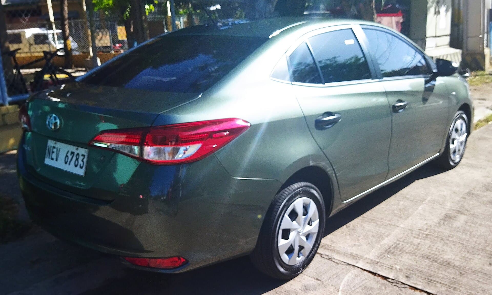 A dark green Toyota Vios sedan seen from the right rear on a concrete driveway, Philippine number plate, dappled shade from trees overhead.
